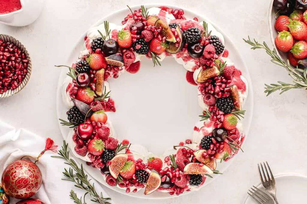 A festive wreath-shaped dessert topped with various berries, figs, cherries, and rosemary sprigs on a white plate.