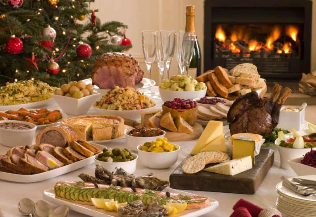 A festive holiday feast graces the table, showcasing roasted meats, assorted cheeses, breads, seafood, and sides. Avoid holiday meal mistakes costing money by planning ahead. A decorated Christmas tree and a lit fireplace provide a cozy backdrop.