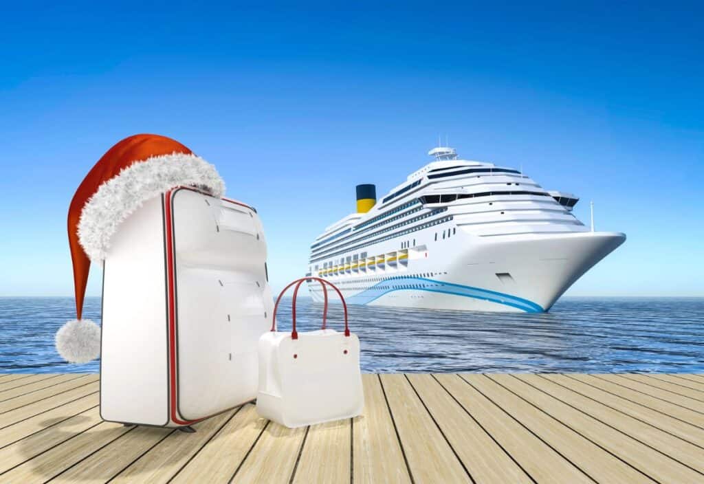 Two suitcases, one with a Santa hat, on a wooden deck with an ocean and cruise ship in the background under a clear blue sky.