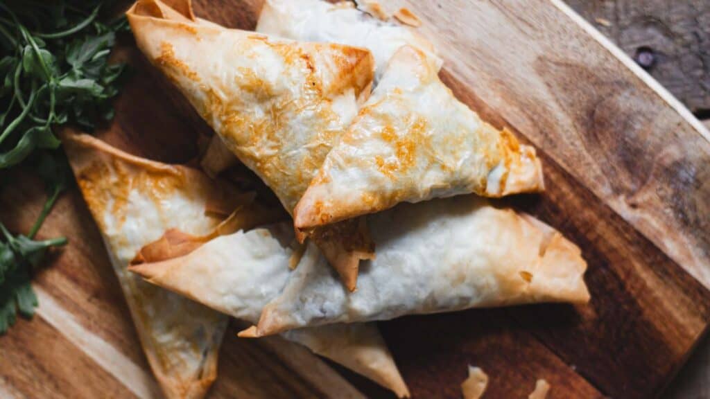 Triangular pastries with a crispy golden crust on a wooden board, garnished with fresh herbs.