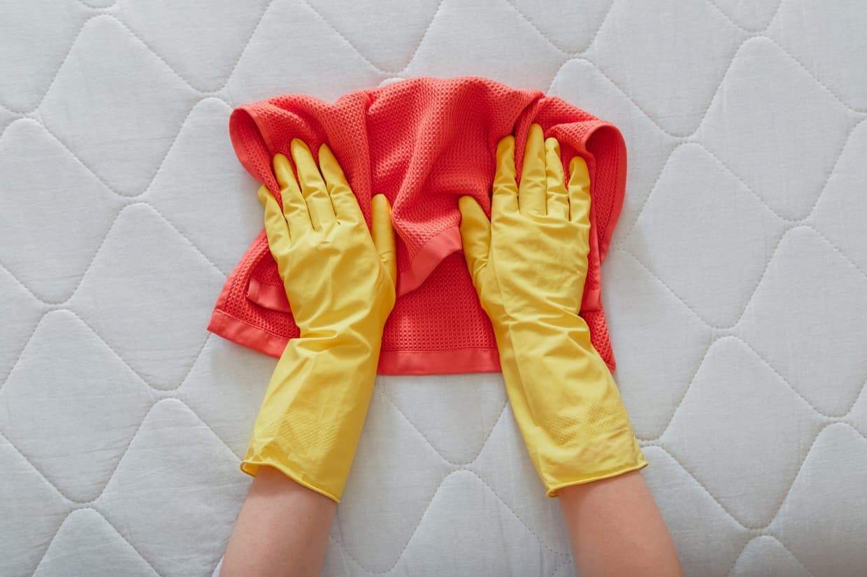 Hands in yellow gloves expertly demonstrate how to clean a mattress, pressing a red cloth against the quilted white surface.