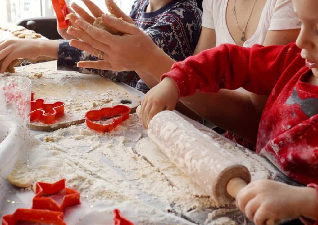 Children and adults making cookies with dough, cookie cutters, and a rolling pin on a floured table.