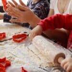 Children and adults making cookies with dough, cookie cutters, and a rolling pin on a floured table.