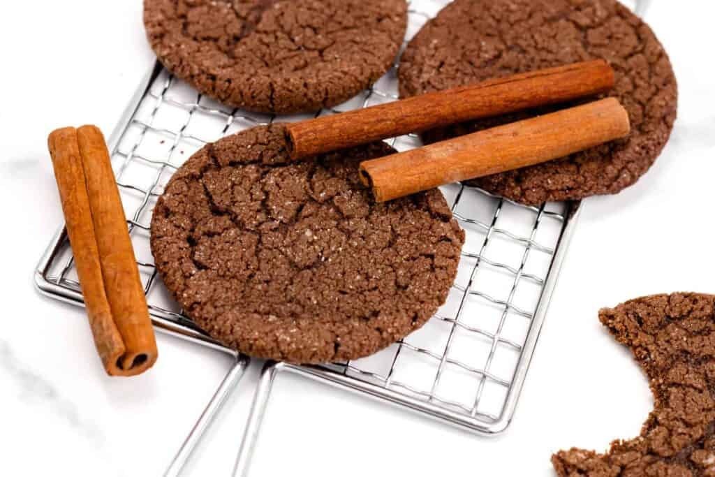 Three gingersnap cookies and two cinnamon sticks on a cooling rack, with one cookie slightly bitten.