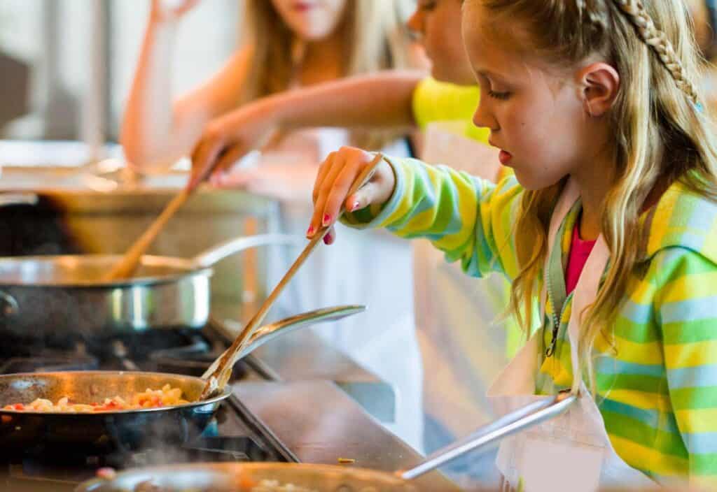 Young girl cooking at a stove, stirring food in a pan with a wooden spoon. Others are seen cooking in the background.