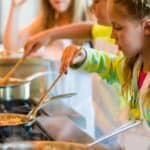 Young girl cooking at a stove, stirring food in a pan with a wooden spoon. Others are seen cooking in the background.