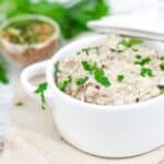 A white bowl filled with creamy spread, garnished with chopped parsley. A spice mix and fresh herbs are in the background.