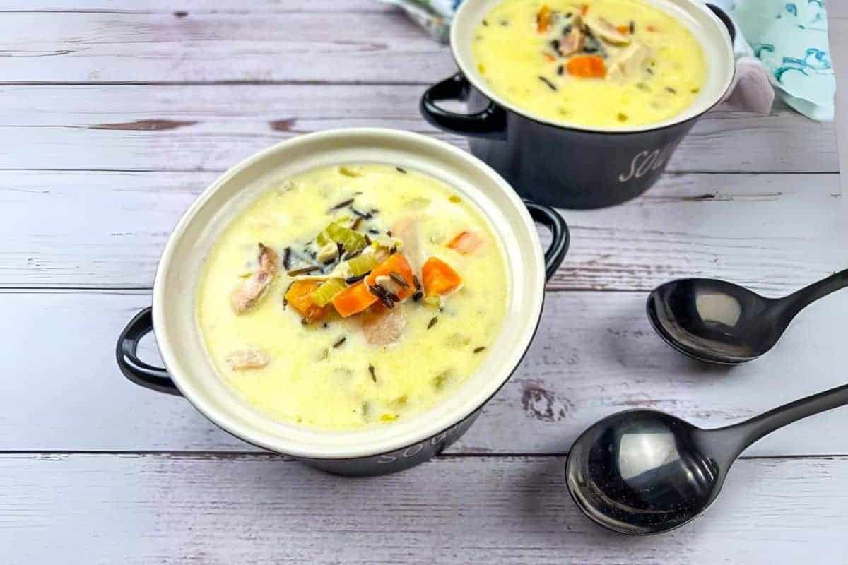 Two bowls of creamy turkey and wild rice soup on a wooden table with two black spoons beside them.