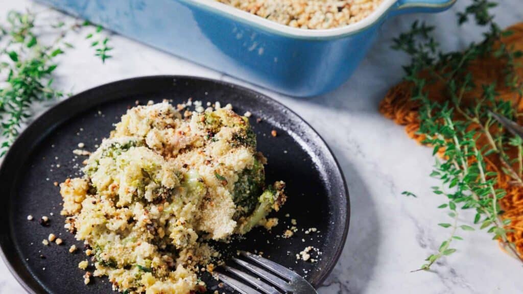 A serving of broccoli casserole topped with breadcrumbs on a black plate. Nearby, a baking dish and sprigs of fresh herbs are on a marble surface.