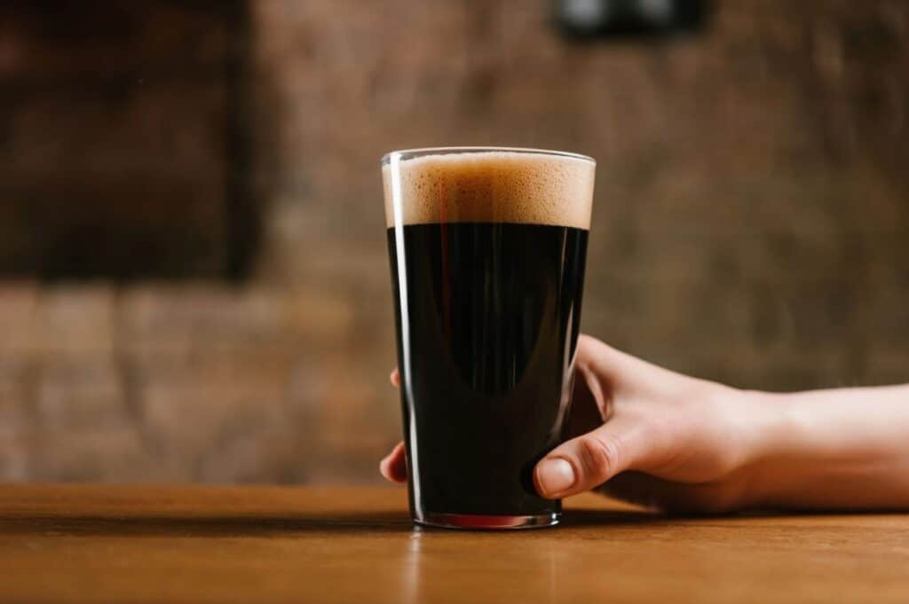 A hand holding a pint glass filled with dark beer, resting on a wooden table with a blurred brown background.