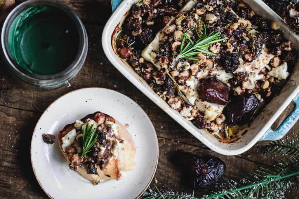 Baked dish with nuts, dried fruits, and herbs in a square pan. A serving on a plate beside a jar with green liquid. Sprigs of rosemary and prunes are scattered on the wooden table.