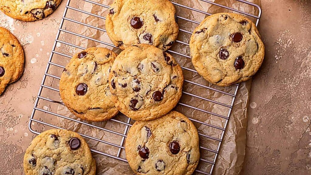 Chocolate chip cookies, made even more delicious by chilling cookie dough beforehand, are cooling on a metal wire rack, placed on brown parchment paper atop a light brown surface.