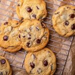 Chocolate chip cookies, made even more delicious by chilling cookie dough beforehand, are cooling on a metal wire rack, placed on brown parchment paper atop a light brown surface.