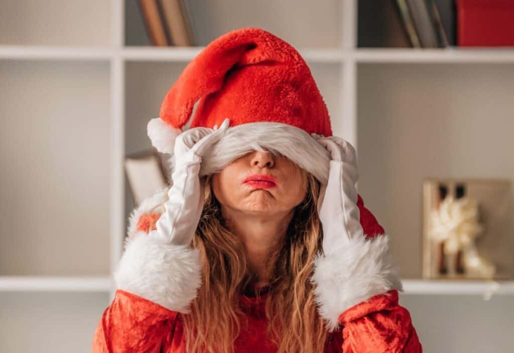 Person in a Santa costume with a hat covering their eyes, wearing white gloves, and standing in front of a white shelf.