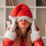 Person in a Santa costume with a hat covering their eyes, wearing white gloves, and standing in front of a white shelf.