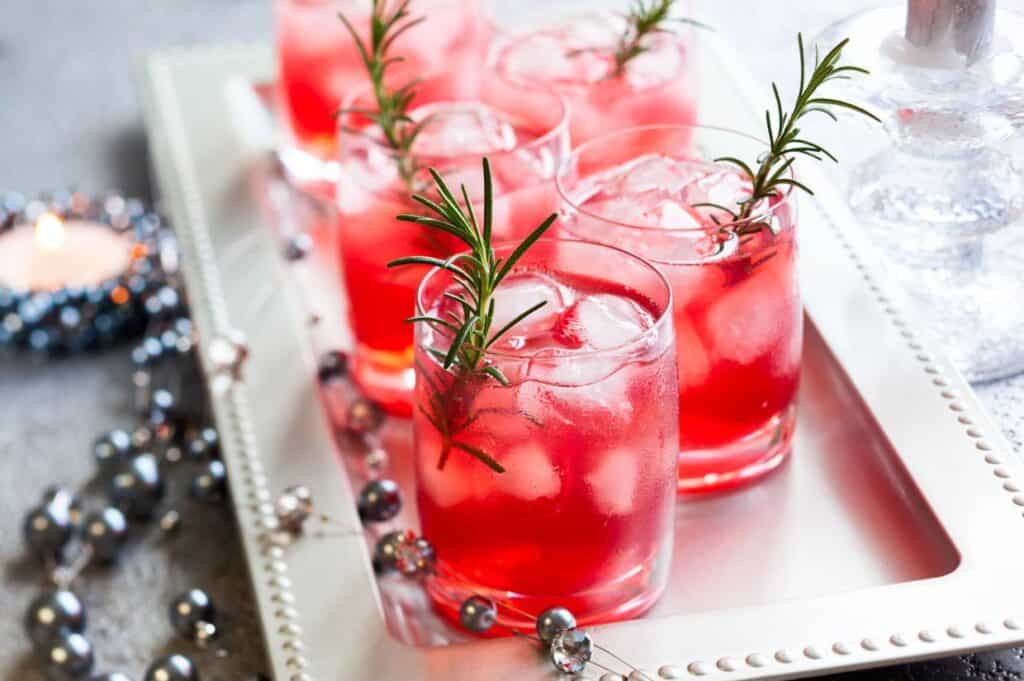 Glasses of red cocktails with ice and rosemary sprigs on a silver tray, next to decorative beads and a lit candle.