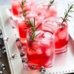 Glasses of red cocktails with ice and rosemary sprigs on a silver tray, next to decorative beads and a lit candle.