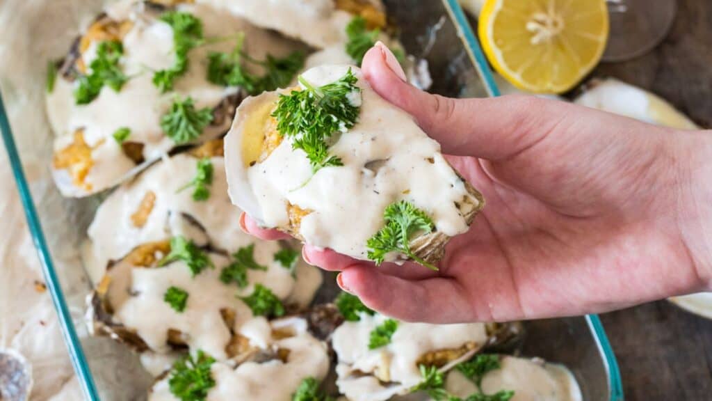 Hand holding a baked stuffed clam topped with creamy sauce and parsley, with a dish of similarly prepared clams and lemon slices in the background.