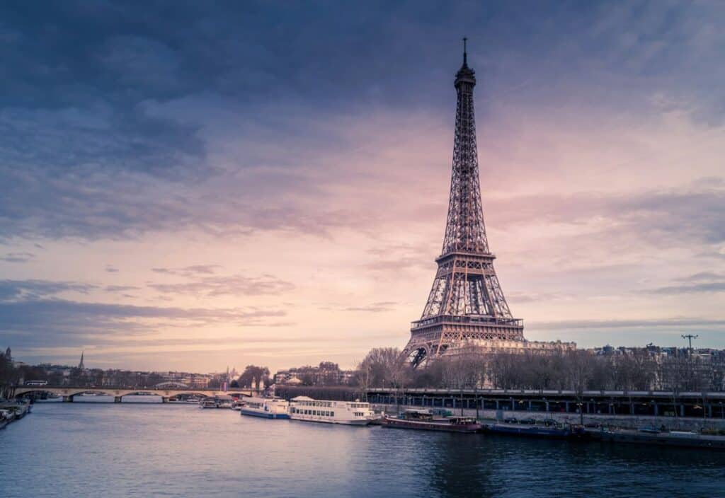 The Eiffel Tower stands by the Seine River under a cloudy sky at dusk, with boats docked along the riverbank.