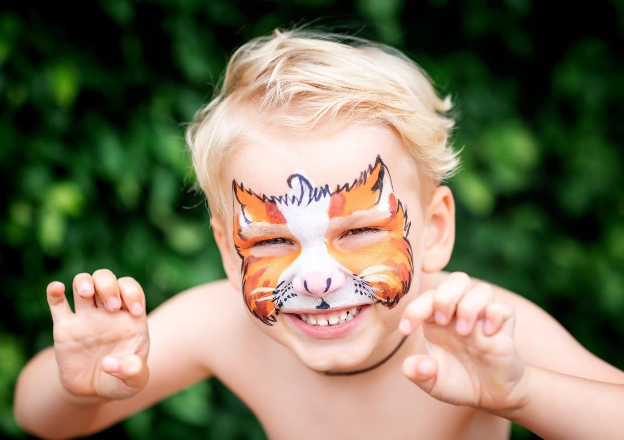 Child with a painted cat face smiles, with hands raised like claws, in front of a leafy background.