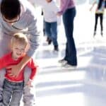 An adult helps a child skate on an ice rink while others skate in the background.