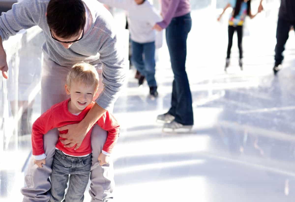 An adult helps a child skate on an ice rink while others skate in the background.