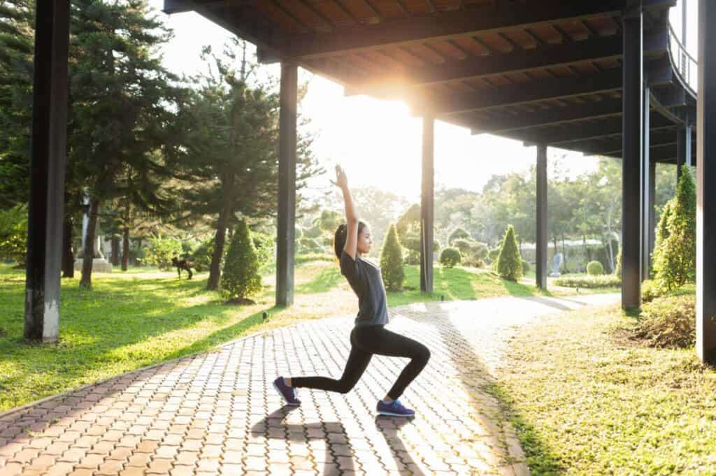 A person performs a yoga pose on a walkway beneath a structure, with sunlight streaming through trees in the background.