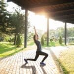 A person performs a yoga pose on a walkway beneath a structure, with sunlight streaming through trees in the background.