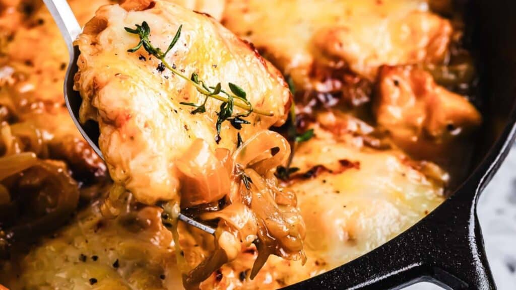 Close-up of a spoon scooping cheesy French onion chicken casserole from a skillet, topped with melted cheese and herbs.