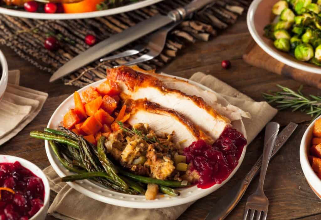 A plate of Thanksgiving food featuring turkey slices, stuffing, green beans, cranberry sauce, and roasted sweet potatoes on a wooden table.