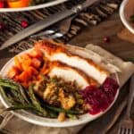 A plate of Thanksgiving food featuring turkey slices, stuffing, green beans, cranberry sauce, and roasted sweet potatoes on a wooden table.