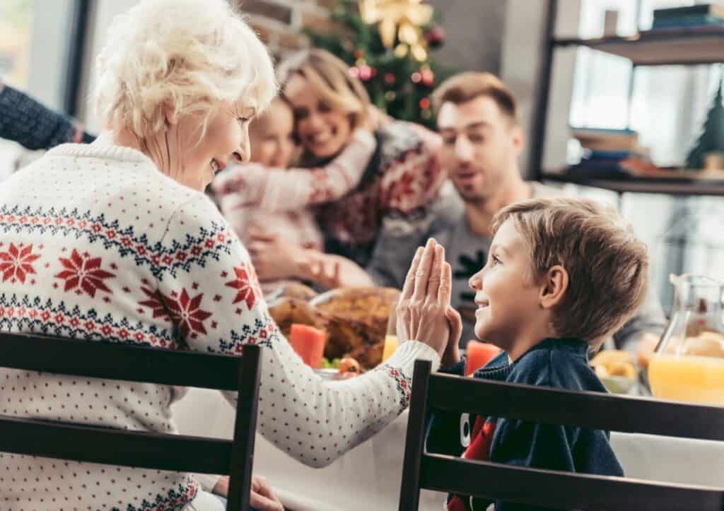 An elderly woman and a young boy high-five at a festive table. Family members are in the background, smiling and interacting.