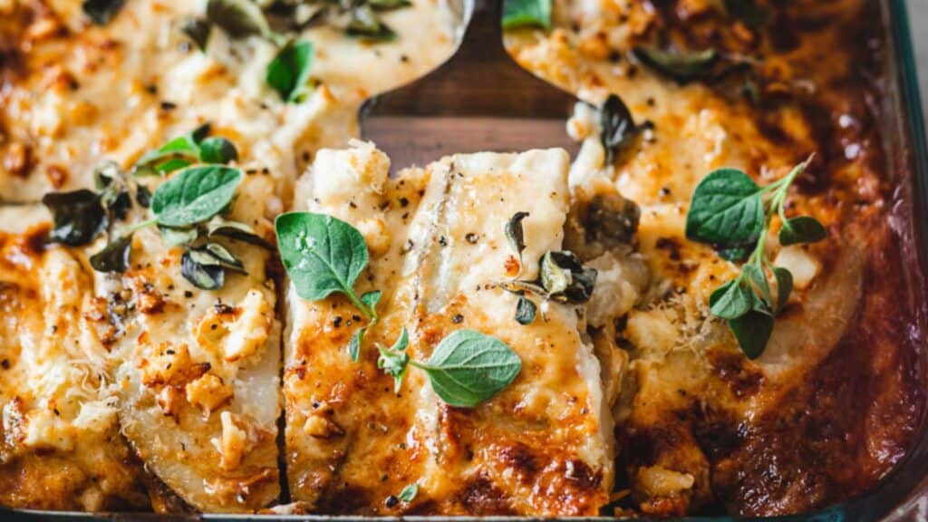 Close-up of a cheesy baked casserole topped with fresh green herbs, featuring browned edges and a portion removed, revealing creamy layers inside.