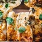 Close-up of a cheesy baked casserole topped with fresh green herbs, featuring browned edges and a portion removed, revealing creamy layers inside.