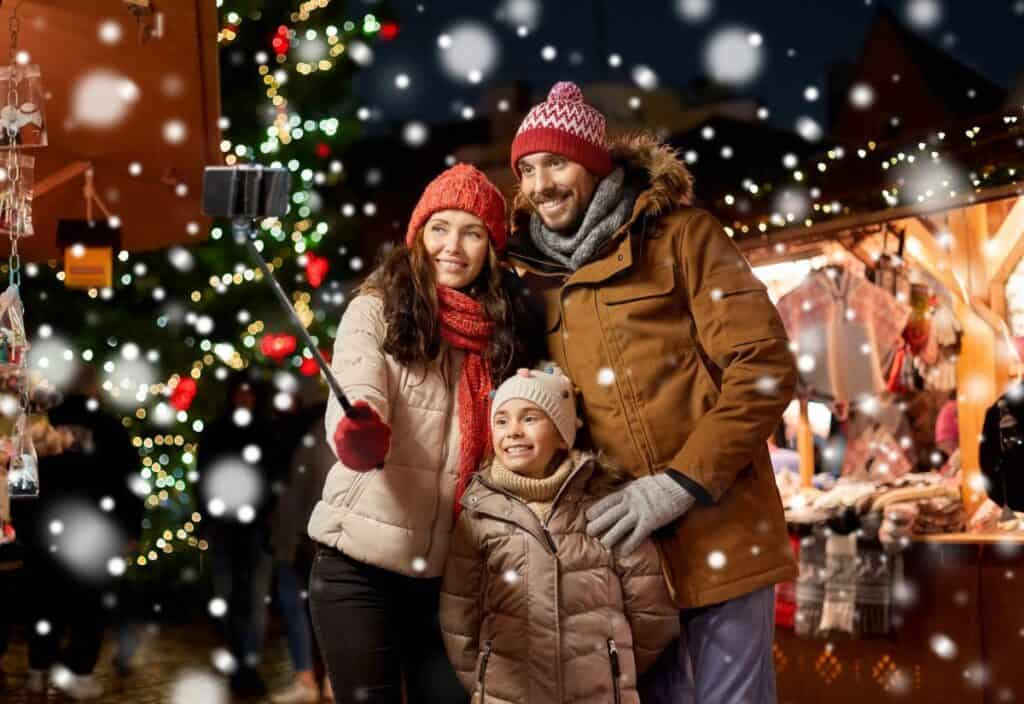 A family of three, bundled in winter clothing, takes a selfie at a festive outdoor market with lights and a Christmas tree in the background, as snow falls gently around them.