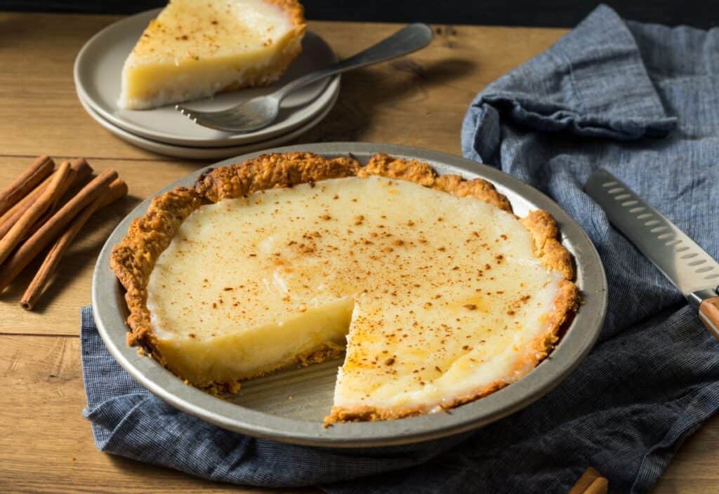 A partially sliced custard pie in a baking dish graces the wooden table, accompanied by cinnamon sticks and a knife. A small plate with pie slices, reminiscent of beloved Thanksgiving regional dishes, sits invitingly in the background.