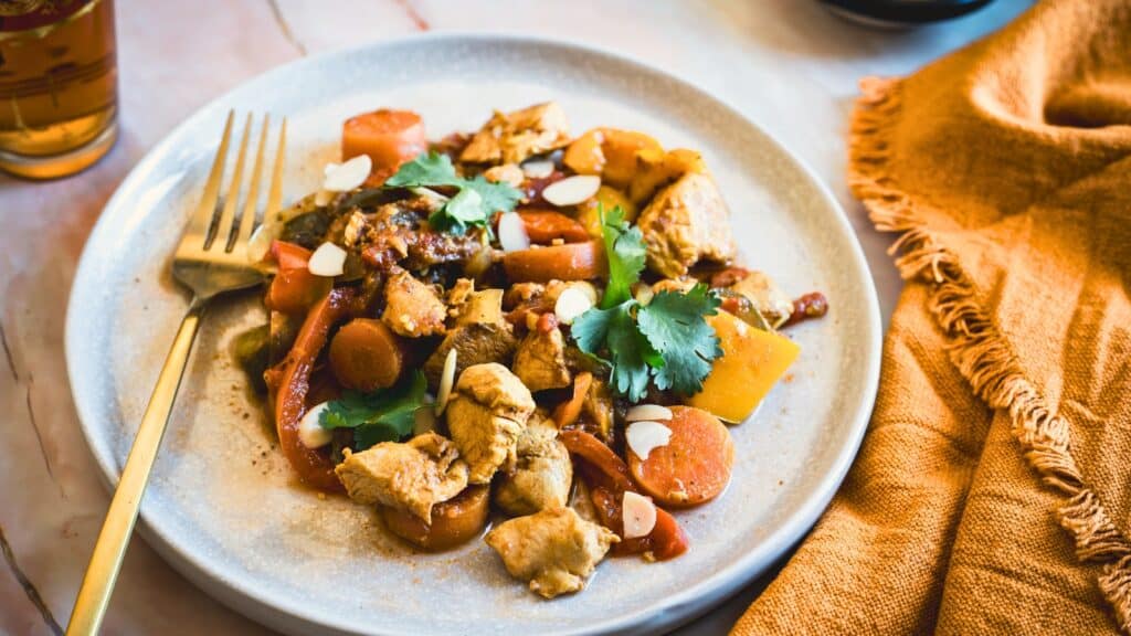 A plate of chicken stew with carrots, peppers, and garnished with cilantro and almond slices, next to an orange napkin and fork.