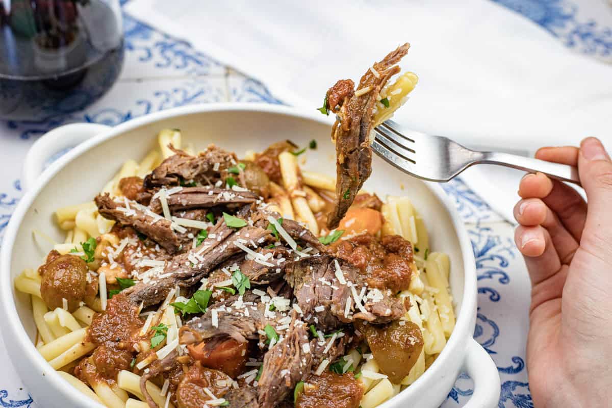 A hand holds a fork with Italian Pot Roast Pasta in a white bowl, topped with grated cheese and herbs.