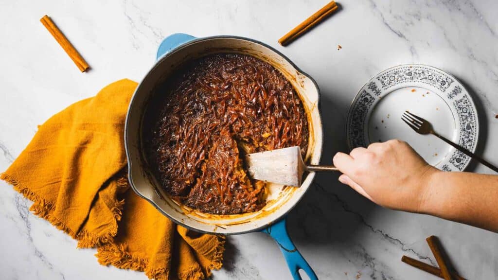 A hand using a spatula to scoop a portion of cooked shredded food from a pot. Nearby are a plate, fork, mustard-colored cloth, and three cinnamon sticks on a marble surface.