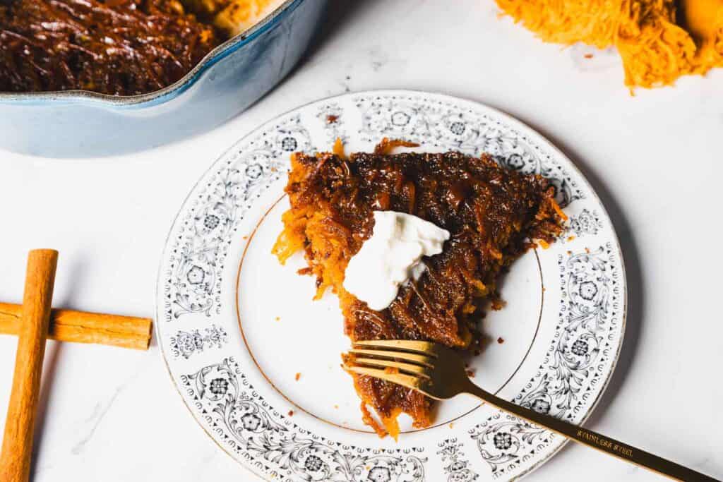 A slice of dessert on a decorative plate with a dollop of cream on top. A gold fork rests on the plate. Nearby are a cinnamon stick and a piece of honeysuckle.