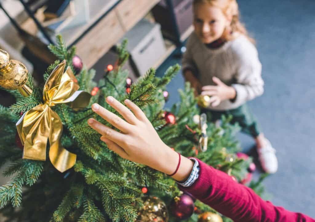 Two children decorate a Christmas tree with ornaments and ribbons. One child reaches for an ornament while the other looks on.