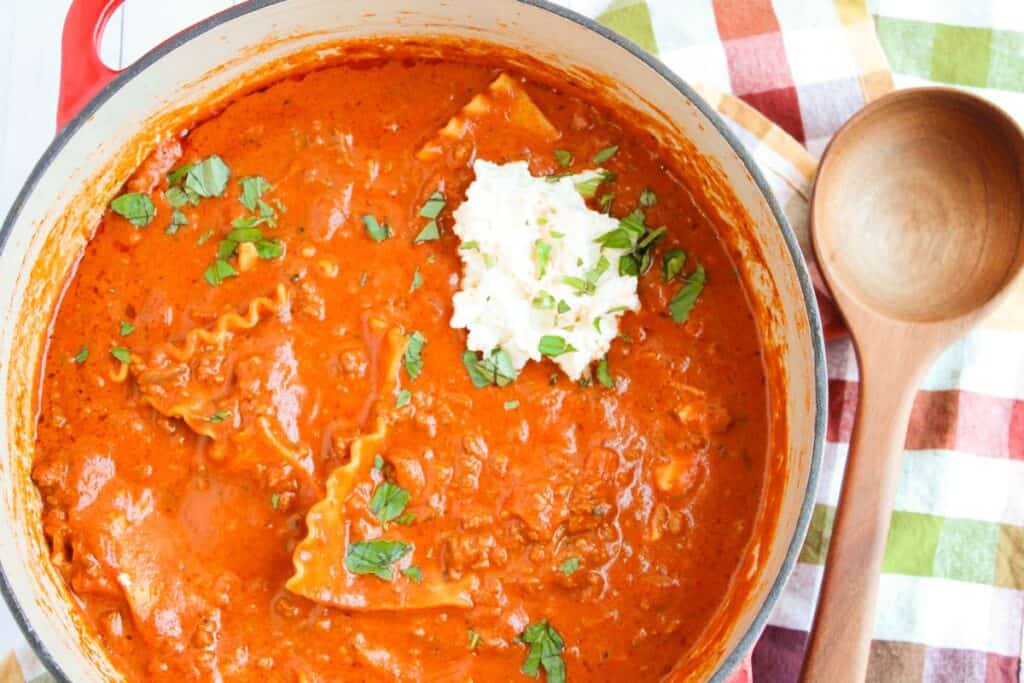 A pot of lasagna soup with lasagna noodles, a dollop of ricotta cheese, and garnished with chopped herbs. A wooden spoon lies beside the pot on a checkered cloth.