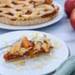 Slice of pumpkin pie with whipped cream and honey on a white plate, with a whole pie and apples in the background.