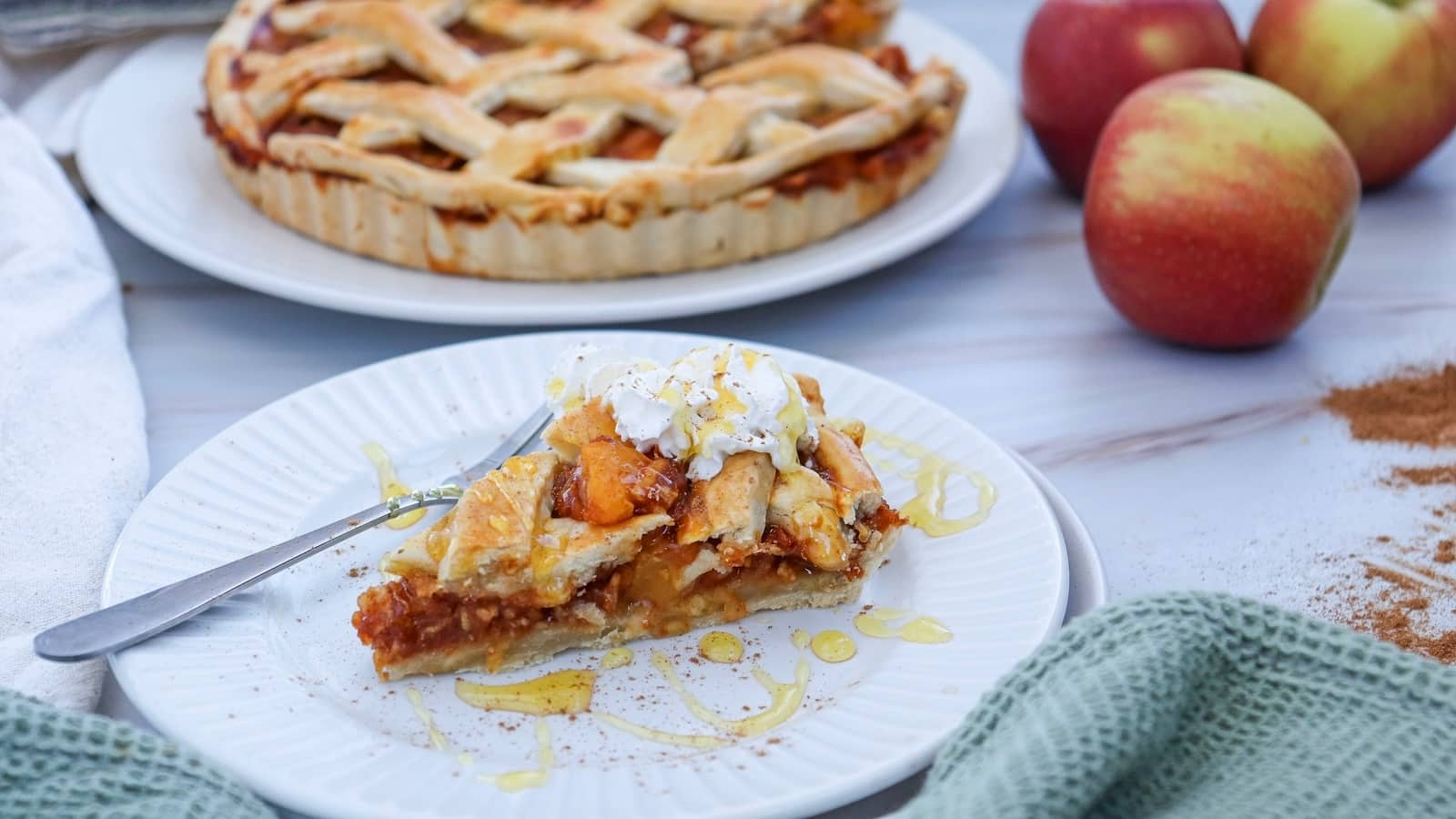 Slice of pumpkin pie with whipped cream and honey on a white plate, with a whole pie and apples in the background.