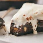 A slice of chocolate brownie with nuts, topped with melting ice cream, sits on a plate next to a spoon.