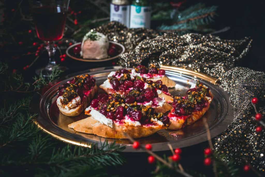 A festive platter with bruschetta topped with ricotta, cranberries, and herbs on a gold tray, surrounded by holiday decorations.