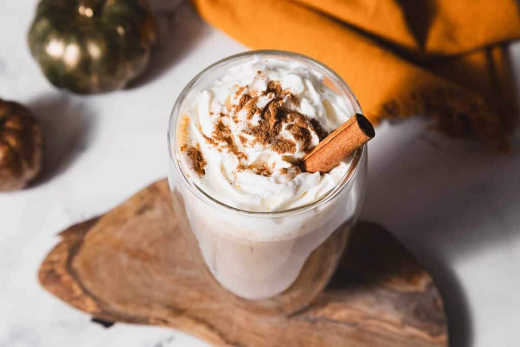 A glass of whipped cream-topped latte with cinnamon and a cinnamon stick on a wooden board. A pumpkin and orange cloth are in the background.