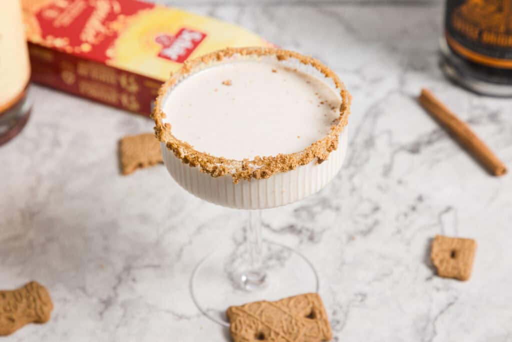 A creamy cocktail in a glass with a crumb-coated rim is surrounded by scattered cookies and a cinnamon stick on a marble surface.