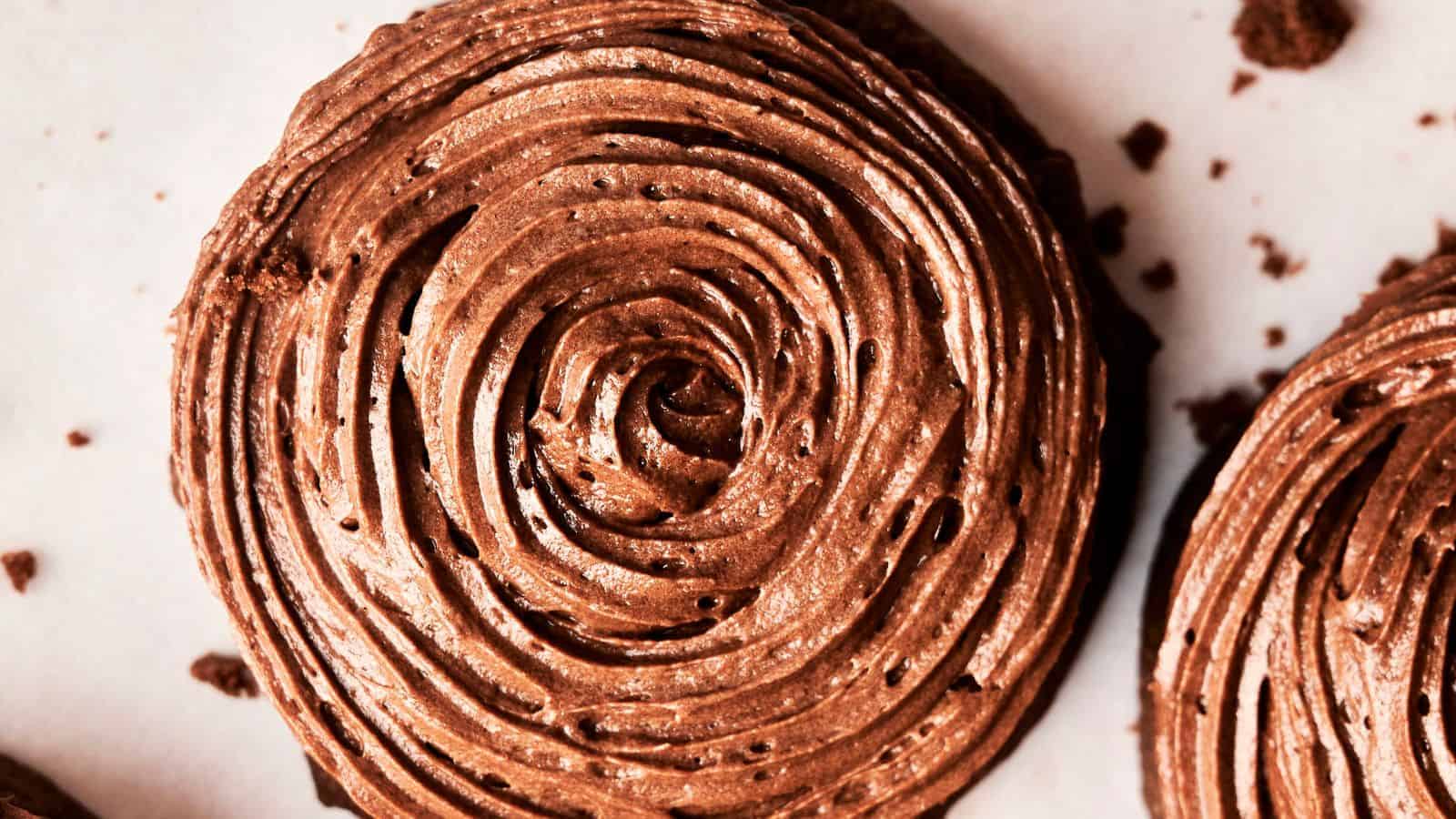 A close-up of a chocolate frosted cupcake with swirl icing on a white surface, reminiscent of Copycat Crumbl Chocolate Cake Cookies and surrounded by crumbs.