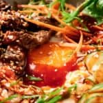 A bowl of bibimbap with sliced beef, rice, sliced vegetables, sesame seeds, and a red sauce.